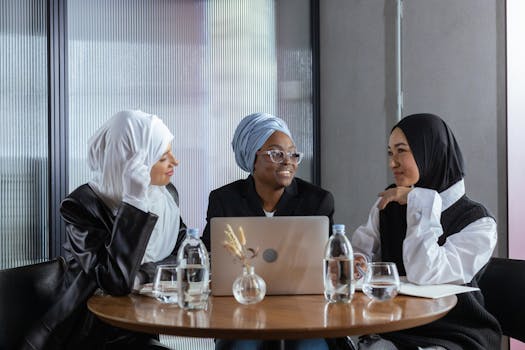 Three diverse women in hijabs discuss ideas around a table with a laptop, promoting inclusivity.